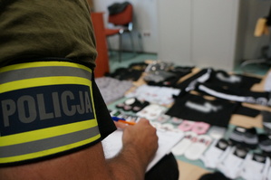 Zbliżenie na napis policja na opasce odblaskowej znajdującej się na ramieniu policjanta nieumundurowanego. W tle widać zamazaną odzież z podrobionymi znakami towarowymi znanych luksusowych marek modowych.