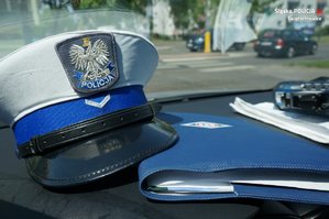 Zbliżenie na czapkę policyjną funkcjonarusza ruchu drogowego leżącą na podszybiu radiowozu, obok notatnik służbowy. W tle widać pojazdy przejeźdzające w okolicy przejścia dla pieszych.