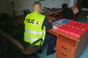 Na zdjęciu nieumundurowany policjant w kamizelce odblaskowej z napisem policja. Przed nim widać na stole zapebezpieczone paczki z papierosami bez polskich znakow akcyzy.