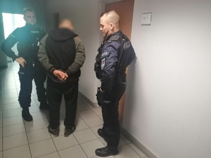 Na zdjęciu dwóch umundurowanych policjantów wraz z zatrzymanym, który na rękach trzymanych z tyłu ma założone kajdanki