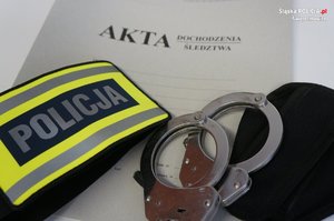 zdjęcie przedstawia: kajdanki, opaska z napisem policja leżące na aktach dochodzeniowo śledczych