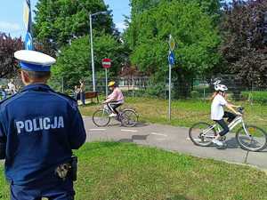 Policjant umundurowany podczas egzaminowania dzieci na kartę rowerową. Dzieci jeżdżą na rowerach w Miasteczku Drogowym.