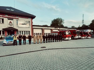 Zdjęcie przedstawia: policjantów i strażaków stojących przy radiowozach