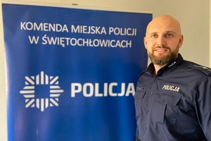 młodszy aspirant Rafał Kamiński na tle niebieskiego banera z napisem Policja i Komenda Miejska Policji w Świętochłowicach