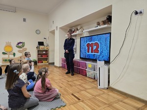 zdjęcie przedstawia policjantkę w przedszkolu i dzieci siedzące na macie, w tle na ekranie wyświetla się numer 112