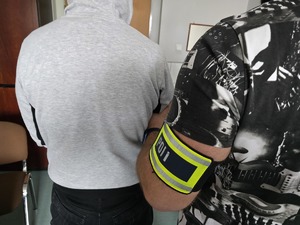 zdjęcie przedstawia policjanta z opaską z napisem policja trzymającego osobę zatrzymaną
