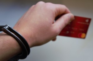 zdjęcie przedstawia rękę w kajdankach trzymającą kartę bankomatową