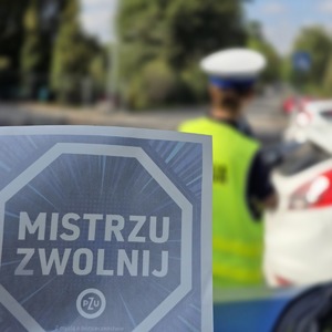 zdjęcie przedstawia: naklejkę z napisem mistrzu zwolnij a w tle policjantka ruchu drogowego