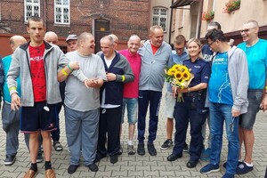 Grupa uczestników spotkania pozuje do wspólnego zdjęcia z policjantką trzymającą bukiet słoneczników przed budynkiem.