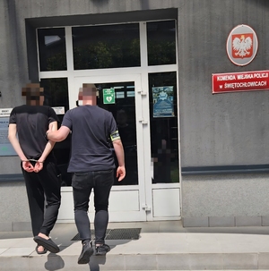 zdjęcie przedstawia policjanta prowadzącego mężczyznę z założonymi kajdankami