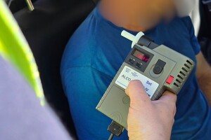 zdjęcie przedstawia policjanta trzymającego alkomat a w tle mężczyznę siedzącego w radiowozie