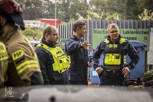 zdjęcie przedstawia policjantów i strażaków stojących przy wraku samochodu i omawiających ćwiczenie