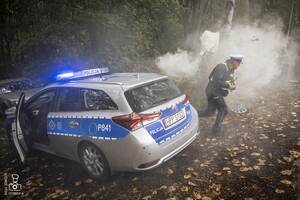 zdjęcie przedstawia wrak z unoszącym się dymem spod maski i biegnący policjant